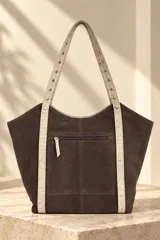 Cartera tipo tote de gamuza color azul marino, con asas dobles de cuero marrón texturado y detalles de tachas metálicas plateadas. Cuenta con una placa metálica frontal con el logo de la marca Pecarí.