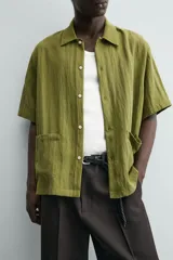 Camisa de corte relajado confeccionada en tejido fluido de lyocell y viscosa, color verde oliva. Presenta cuello solapa, manga corta, cierre frontal de botones y dos bolsillos plastrón en la parte inferior.