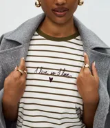 Remera de corte boxy fit, con diseño a rayas horizontales en color blanco y verde oliva. Presenta cuello redondo en contraste, mangas cortas y un bordado frontal con la frase 'I live so I love' acompañada de un pequeño corazón.