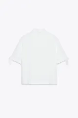 Camisa de manga corta confeccionada en tejido de ramio color blanco. Posee cuello solapa, cierre frontal con botones y detalle de lazos en el acabado de las mangas.