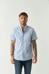 Camisa de manga corta con estampado de rayas verticales finas en color celeste y blanco. Presenta cuello abotonado, cierre frontal con botones y un bolsillo en el pecho con logo bordado.