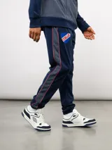 Pantalón deportivo de piqué color azul marino, con franjas laterales en contraste rojo y blanco. Presenta el escudo del Club Nacional de Football bordado en la pierna derecha y una etiqueta conmemorativa en la izquierda.