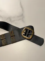 Cinto faja de cuero color suela con hebilla metálica cuadrada y apliques de varillas metálicas verticales en la estructura. Presenta un diseño artesanal con logo grabado en el extremo.