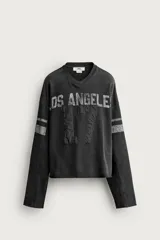 Sudadera cropped fit de color negro con efecto lavado vintage, cuello en V y manga larga. Presenta estampado con texto "LOS ANGELES" y el número "17" en parches acolchados en el frente, además de bandas grises en las mangas.