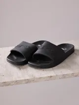 Ojotas tipo slide color negro, con banda ancha superior que presenta un diseño gráfico en relieve con el logo de la marca. Suela anatómica de PVC con textura antideslizante.