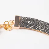 Collar tipo gargantilla rígida de diseño tubular, recubierto con cristales brillantes en tono gris oscuro, con cierre y cadena de extensión en metal dorado.