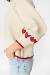 Sweater de punto color crudo con diseño de corazones rojos en las mangas y detalles de ribete rojo en cuello, puños y ruedo. Presenta un corte holgado con mangas abullonadas.