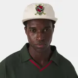 Gorra color beige con visera plana y parche bordado en el frente con escudo y la inscripción 'GOLF'.
