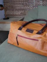 Bolso tipo weekender de cuero marrón, con asas de mano y bolsillo frontal con cierre metálico.