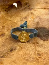 Pulsera con dije redondo dorado con diseño abstracto y cinta de terciopelo elástico color gris claro.