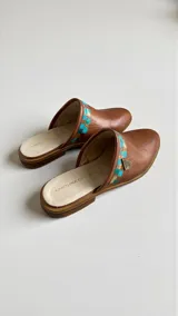 Sandalias tipo mules color marrón terracota, confeccionadas en cuero, con punta redonda, suela plana y detalles pintados a mano en tonos turquesa y dorado con aplique metálico triangular en el lateral.