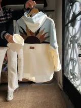 Campera de estilo oversize confeccionada en una combinación de corderito blanco y denim celeste. Presenta un diseño frontal con un aplique bordado de sol en tonos tierra y una etiqueta de marca en el centro. Posee mangas largas y cuello alto.