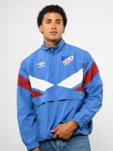 Buzo deportivo de nylon con medio cierre frontal y cuello alto. Presenta un diseño de bloques de color en azul, blanco y bordó, con el escudo del Club Nacional de Football y el logo de Umbro en el pecho.