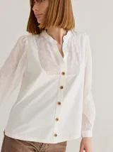 Camisa de corte recto y manga larga, confeccionada en algodón blanco. Presenta un canesú superior con bordado inglés floral y detalle de placa metálica con logo en la espalda.