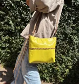 Cartera bandolera de cuero flexible color amarillo, con trenza artesanal al frente, interior forrado y correa larga con herrajes de metal.