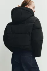 Campera puffer acolchada de color negro, con cuello envolvente, capucha integrada, manga larga, bolsillos laterales ocultos y bajo con elástico. Cierre frontal con botones a presión ocultos por solapa.