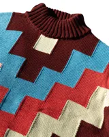 Sweater de cuello alto confeccionado en tejido de punto con diseño geométrico en bloques de colores bordó, rojo, celeste y beige. Presenta puños y ruedo acanalados y un pequeño parche decorativo en el frente.