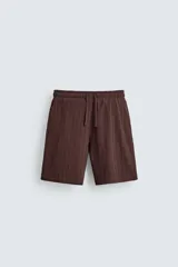 Bermuda de corte relajado color marrón oscuro, confeccionada en tejido jacquard con textura acanalada vertical. Presenta cintura elástica ajustable con cordón, bolsillos delanteros y un bolsillo trasero de plastrón.