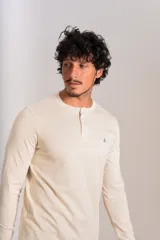 Remera de manga larga tipo Henley color beige, con cuello redondo y cierre frontal parcial con botones. Presenta un pequeño logo bordado en el pecho.