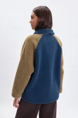 Campera de sherpa con diseño color block, combinando cuerpo azul, mangas marrones y detalles en rosa pálido. Presenta cierre frontal con botones a presión, bolsillo superior con cierre y bolsillos laterales con cremallera.
