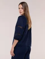 Blusa blanca de calce suelto y caída ligera, con escote en V y cuello Mao pequeño. Presenta mangas tres cuartos abullonadas con elástico en los puños y detalles de calado o puntilla encastrada. Espalda lisa con canesú y leve frunce.