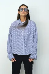 Camisa de corte holgado con estampado de rayas verticales azules y blancas. Presenta cuello clásico, cierre frontal con botones, hombros caídos y mangas largas con puños abotonados. El diseño incluye un canesú con pliegue en la espalda y bajo redondeado con la parte posterior ligeramente más larga.