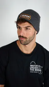 Gorro tipo beanie de punto color gris, con parche frontal bordado con el logo de la marca Duxon en colores blanco y naranja.
