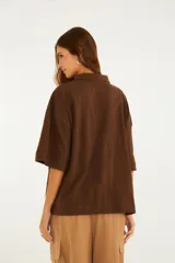 Remera tipo polo marrón, de corte oversize, con cuello clásico, cartera con botones y mangas amplias hasta el codo. Cuenta con un detalle bordado circular en el pecho.