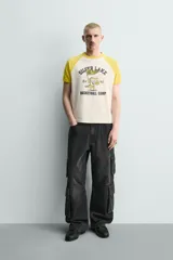 Camiseta de corte relajado con mangas raglán cortas en color amarillo y cuerpo en color crema. Presenta un estampado gráfico frontal con temática de campamento de básquetbol y un cocodrilo.