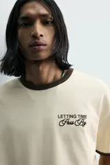 Camiseta de corte regular, color beige con cuello y bordes de mangas en color marrón oscuro. Presenta un diseño de texto bordado en el pecho con la frase 'Letting Time Pass By'.
