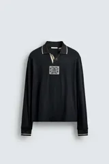 Polo de manga larga en tejido técnico acanalado color negro, con cuello solapa y abertura frontal. Presenta puños con ribete a contraste y una etiqueta aplicada en el pecho con el diseño conmemorativo de la Copa Mundial de la FIFA 1966.