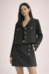 Conjunto de chaqueta crop y falda corta en tejido de tweed negro, con botones metálicos decorativos con pedrería. La chaqueta presenta solapas clásicas, bolsillos frontales con solapa en el pecho y manga larga. La falda presenta una silueta recta con detalle de botonadura lateral.