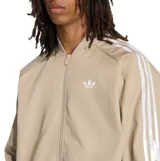 Campera Adidas Adicolor de color beige con cierre frontal y cuello bomber. Cuenta con las clásicas tres tiras blancas en las mangas y el logotipo del trébol bordado en el pecho.