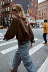 Camisa marrón de manga larga con cuello solapa y cierre frontal de botones. Los puños terminan con lazos anudados.