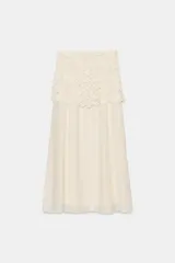 Falda midi de color crudo, con diseño combinado que presenta una parte superior de crochet con motivos florales y una parte inferior de tela fluida con textura arrugada. Incluye forro interior y cierre lateral con cremallera oculta.