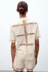 Blusa de manga corta confeccionada en algodón, con cuello clásico y cierre frontal de botones. Presenta un diseño combinado con paneles de tejido bordado floral y detalles de crochet en cuello, mangas, frente y ruedo.