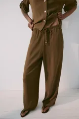 Pantalón de tiro medio color marrón, con cintura elástica y cordón ajustable con abalorios decorativos. Presenta bolsillos laterales y perneras rectas con diseño texturizado.