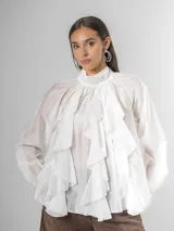Camisa blanca de algodón italiano con cuello alto ajustable mediante lazo trasero, mangas abullonadas y un gran shabot frontal con volados en cascada.