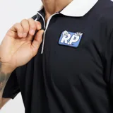 Polo de manga corta color negro con cuello blanco y cierre frontal parcial. Presenta un pequeño parche bordado con un logo azul y blanco en el pecho izquierdo.