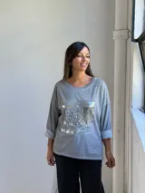 Remera gris de manga larga con cuello redondo y diseño holgado. Presenta un aplique frontal rectangular con detalles de lentejuelas plateadas y estampado tipográfico.