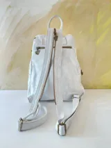 Mochila de cuero blanca con diseño artístico pintado a mano en tonos dorados, marrones y negros en el frente, múltiples bolsillos con cierre y herrajes de metal.