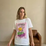 Remera blanca de corte holgado con estampa frontal colorida que incluye rayas verticales rosadas y un diseño gráfico de un trago con rodaja de naranja. Confeccionada en mezcla de algodón y poliéster.