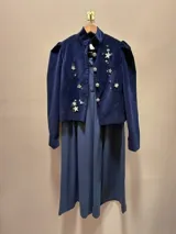 Chaqueta de paño de lana y cashmere color verde oscuro, de corte estructurado con cuello mao, mangas largas con hombros abullonados y puños con tajo. Cuenta con cierre frontal de botones dorados grandes y detalles de bordados dorados artesanales en el pecho y el bajo.