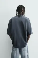 Camiseta de algodón color gris oscuro, corte relajado (relaxed fit), con cuello redondo y manga corta. Presenta dos inscripciones bordadas en hilo blanco en el frente.