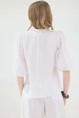 Camisa de lino color beige, de silueta romántica con escote a la base y mini volado. Cuenta con mangas 3/4 abullonadas con frunces y terminación en elástico. Posee detalles de puntillas negras en contraste en el frente y cierre frontal con botones ocultos bajo cartera.