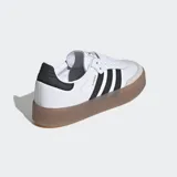 Zapatillas Adidas Sambae blancas con las tres tiras laterales en negro, refuerzo de gamuza gris en la puntera, contrafuerte trasero negro y suela de goma color caramelo.