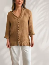Camisa de lino color tostado, de corte relajado, con cuello camisero abierto y cierre frontal de botones oscuros. Presenta mangas largas con puños ligeramente doblados.