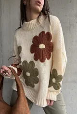 Sweater de tejido de punto color natural, con diseño de flores grandes aplicadas en tonos marrón y verde oliva. Presenta cuello redondo, mangas largas y corte holgado.