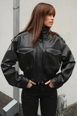 Cazadora de estilo bomber en efecto piel color negro, con cuello subido, manga larga y bajo elástico. Presenta trabillas en los hombros, bolsillos frontales con solapa y cierre de cremallera con botones a presión ocultos.