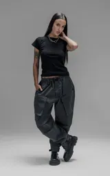 Pantalón de estilo utilitario confeccionado en textil técnico ripstop color gris oscuro. Presenta un corte oversize con cintura elástica, cierre frontal con broche técnico, bolsillos delanteros de gran tamaño con cierre y sistema de ajuste en los tobillos mediante broches snap.